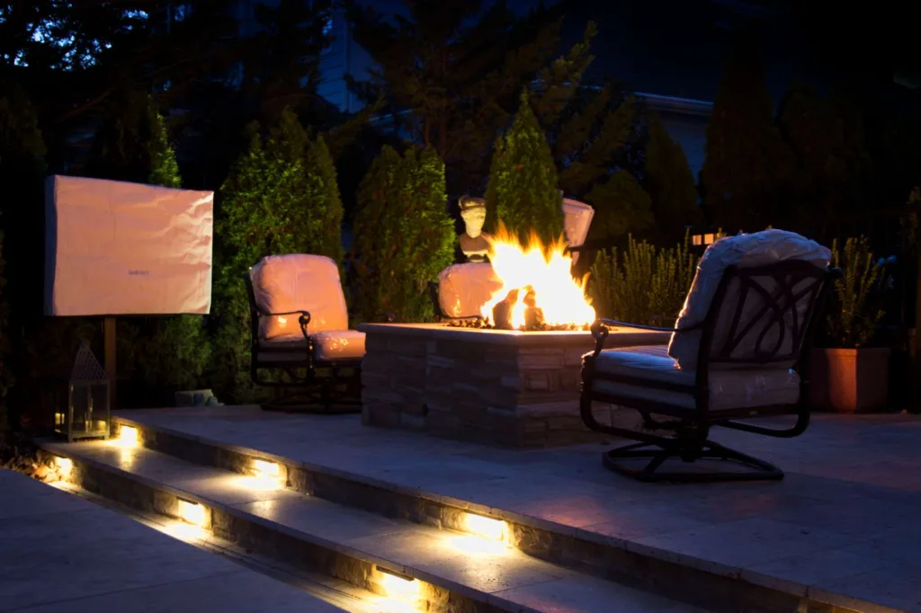 Outdoor area with firepit, chairs, and tv. Everything has weather protectant covers on it. Landscape lighting on surrounding areas and lighting on steps to lounge area.