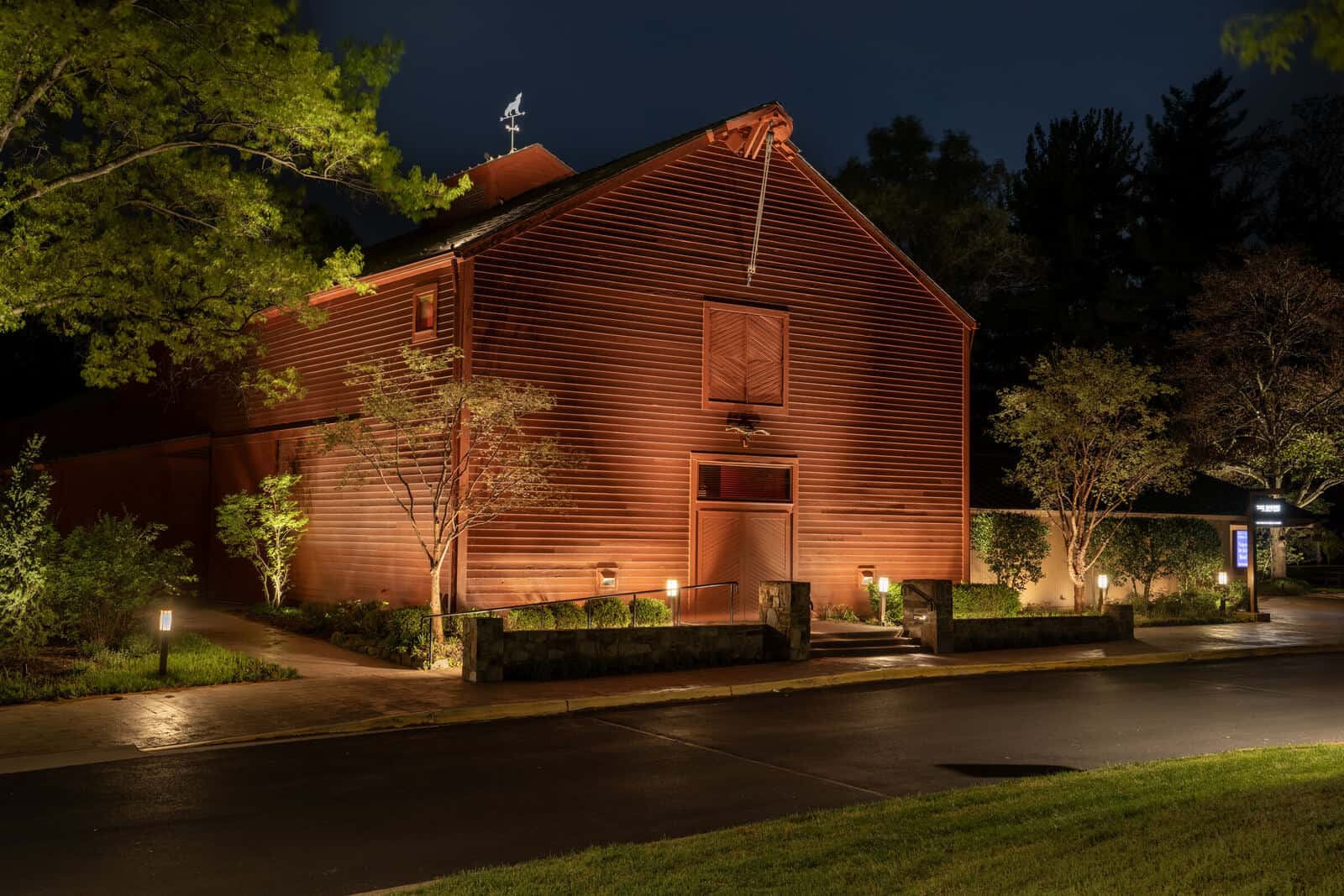 The Barns of Wolf Trap in Vienna, Virginia, at night, the red barn gable washed in warm light with uplit trees and garden beds