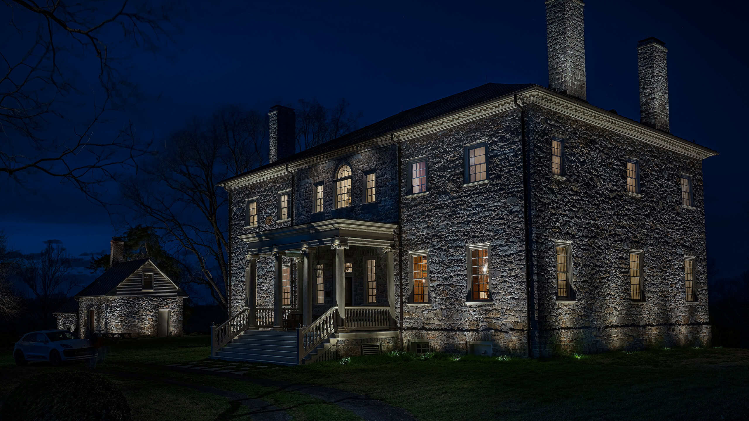 A three-story historic fieldstone manor at night, with soft grazing light on the stone walls and a lit front porch.