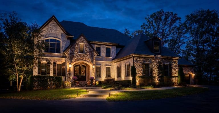 Stone manor home at dusk with soft uplighting on its gables and arched entry, path lights along the walkway and lit trees.