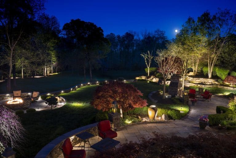 Terraced garden at dusk with uplit trees and a red Japanese maple, lit stone walls, and path lights along a winding walkway.