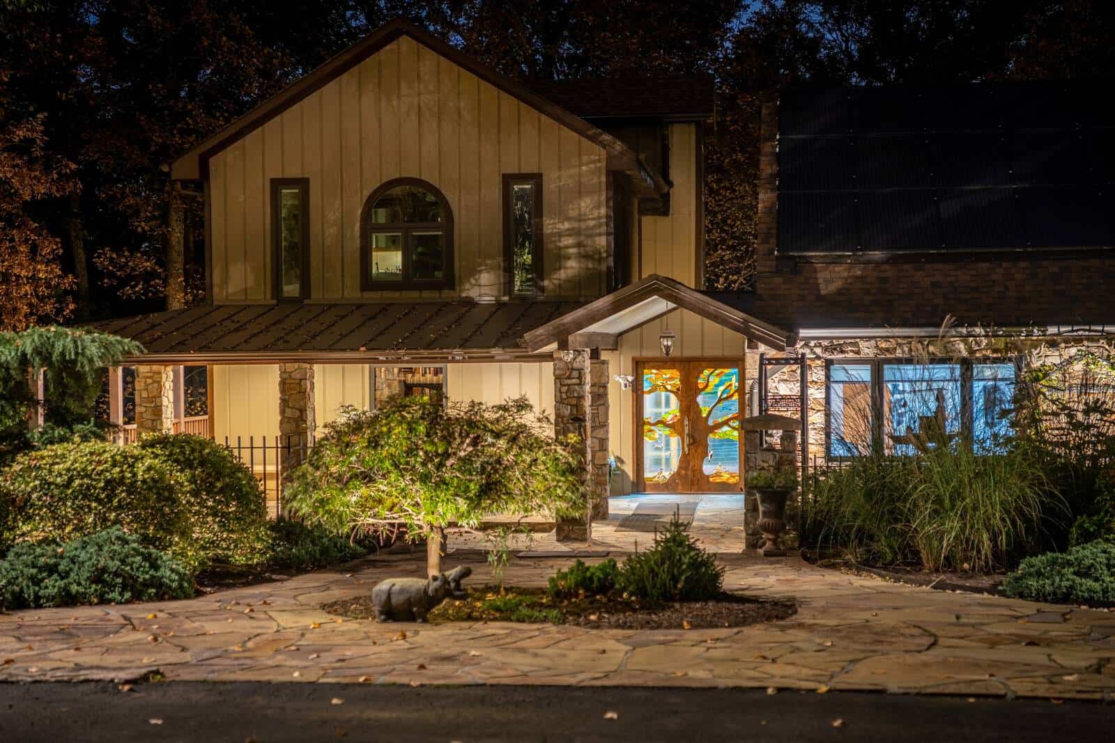 Board-and-batten home at night in Catharpin, Virginia, with a glowing stained-glass front door, uplit Japanese maple, and flagstone entry walk