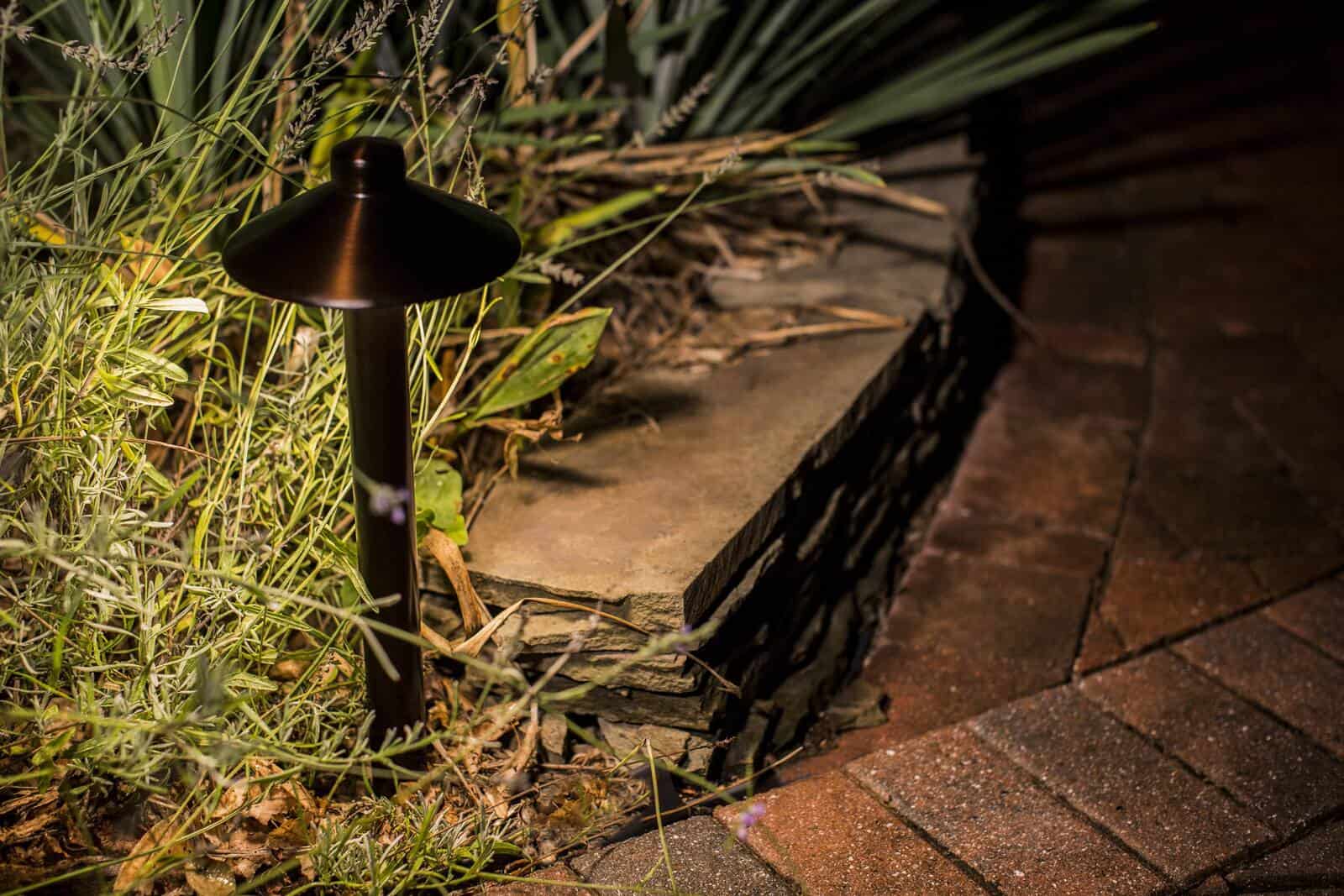 Close-up of a bronze Sterling Lighting path light casting warm, glare-free light beside a stacked stone wall at night