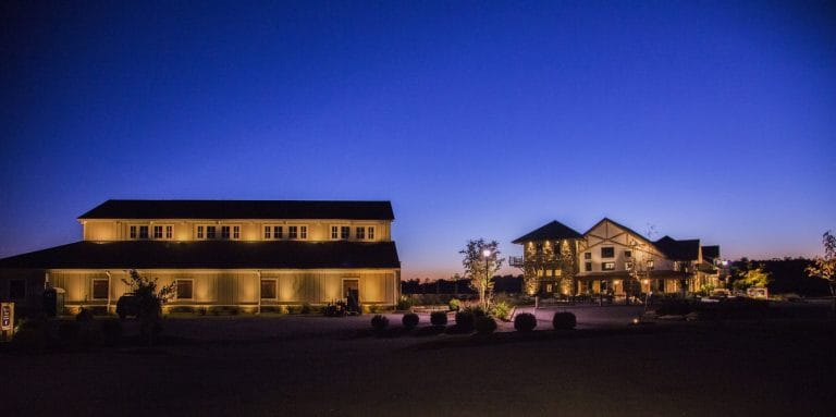Wide view of a hilltop winery at twilight, its long barn and tower lit by warm facade lighting against a deep blue sky.