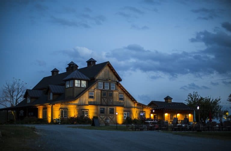 Timber-frame winery barn at dusk with warm uplighting washing the stone base, cupolas and entry under a cloudy blue sky.