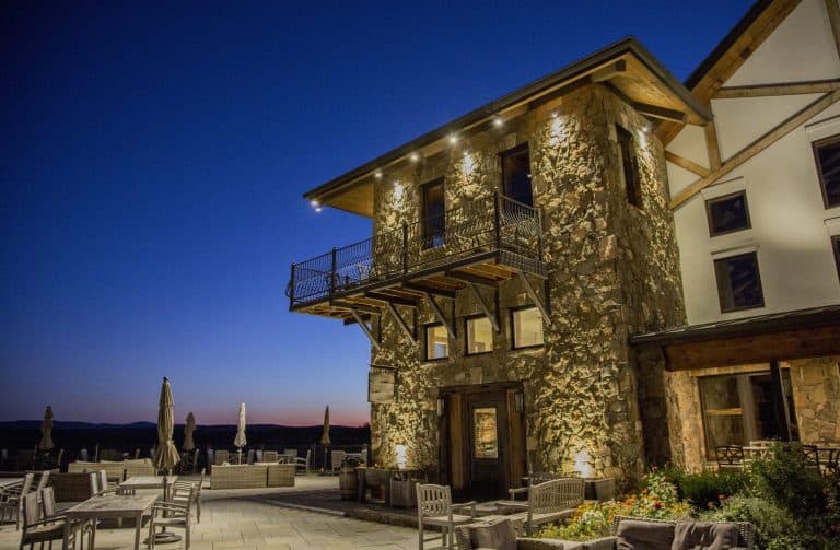 Stone tower with grazing light on its fieldstone walls and timber balcony, rising above a lit terrace with patio seating.