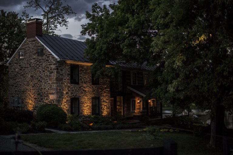 Historic stone farmhouse with warm uplighting on its stone walls and front gardens at dusk in western Loudoun County