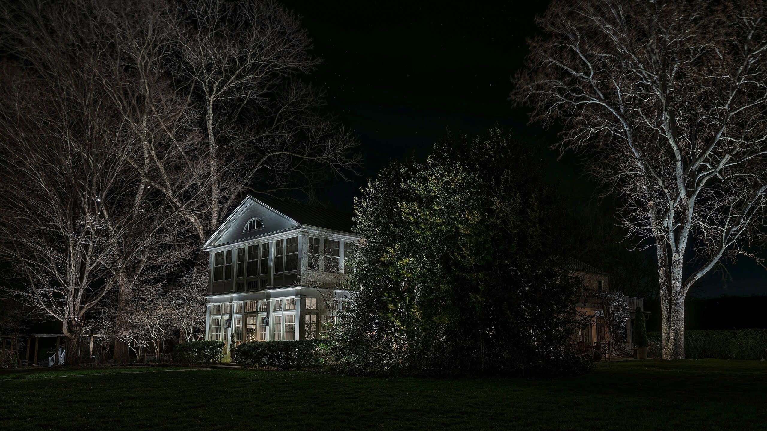 A white frame house with a lit sunporch at night, framed by tall uplit sycamore trees.