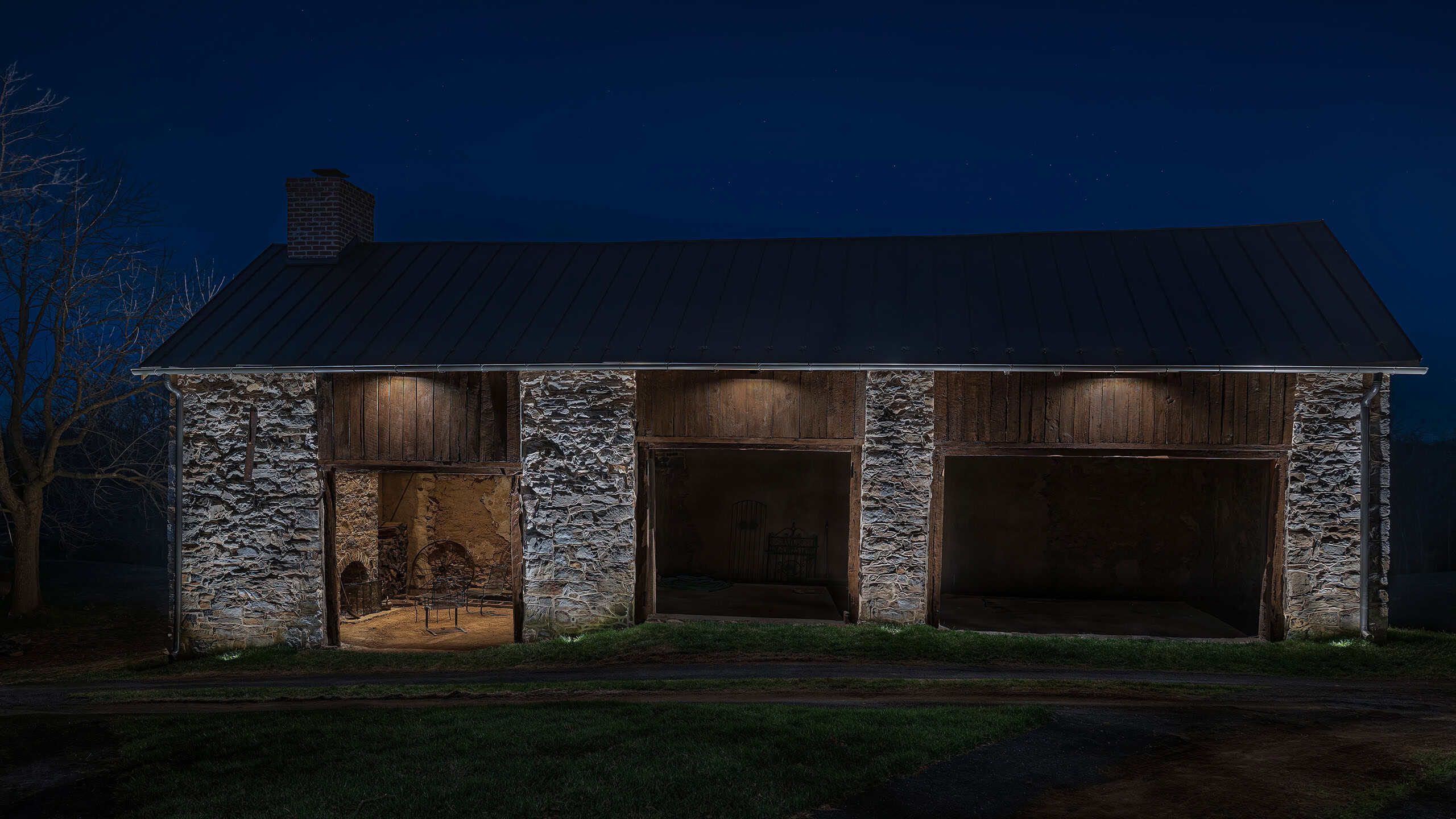 A historic open-bay stone barn at night, with warm light grazing its stone piers and timber bays.