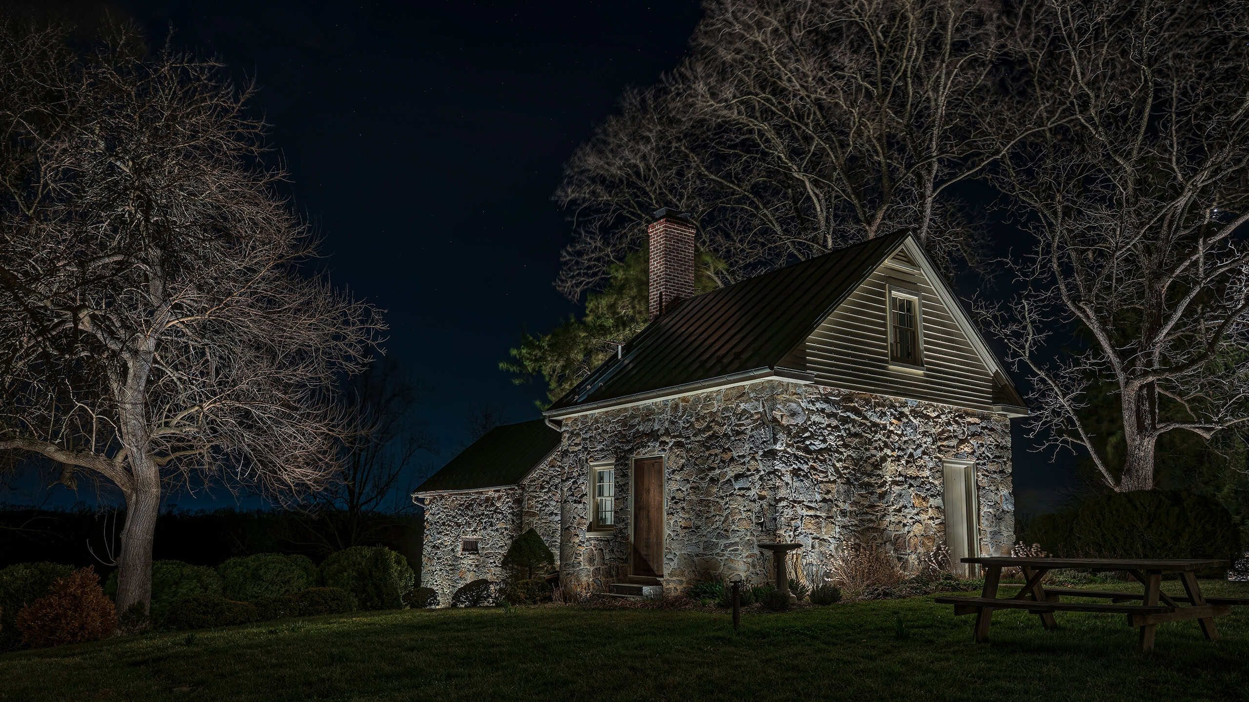 A historic stone cottage at night, with grazing light on its walls and uplit mature trees beside it.
